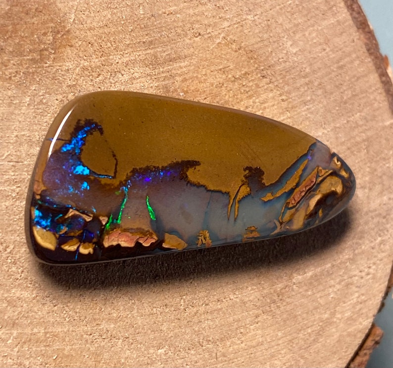 May include: A brown, blue, and green opal cabochon with a rough, natural shape. The opal has a dark brown base with flashes of blue, green, and purple.