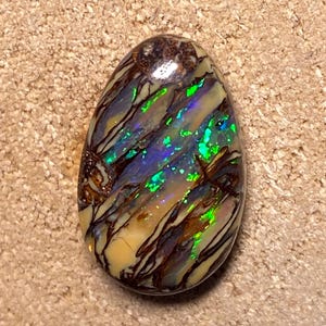 Könnte beinhalten: Ein tropfenförmiger Opal-Cabochon mit einem braunen, weißen und grünen irisierenden Muster. Der Opal hat eine glatte, polierte Oberfläche.