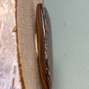 May include: A dark brown and gold opal cabochon with a blue and green iridescent sheen. The opal is set in a dark brown bezel.
