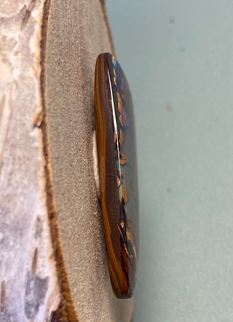 May include: A dark brown and gold opal cabochon with a blue and green iridescent sheen. The opal is set in a dark brown bezel.