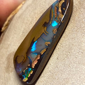 May include: A dark brown, green, and blue opal gemstone with a teardrop shape. The opal has a high polish and is set in a dark brown backing.