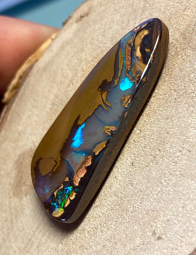 May include: A dark brown, green, and blue opal gemstone with a teardrop shape. The opal has a high polish and is set in a dark brown backing.