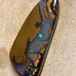 May include: A dark brown, green, and blue opal cabochon with a teardrop shape. The opal has a high polish and shows flashes of color.