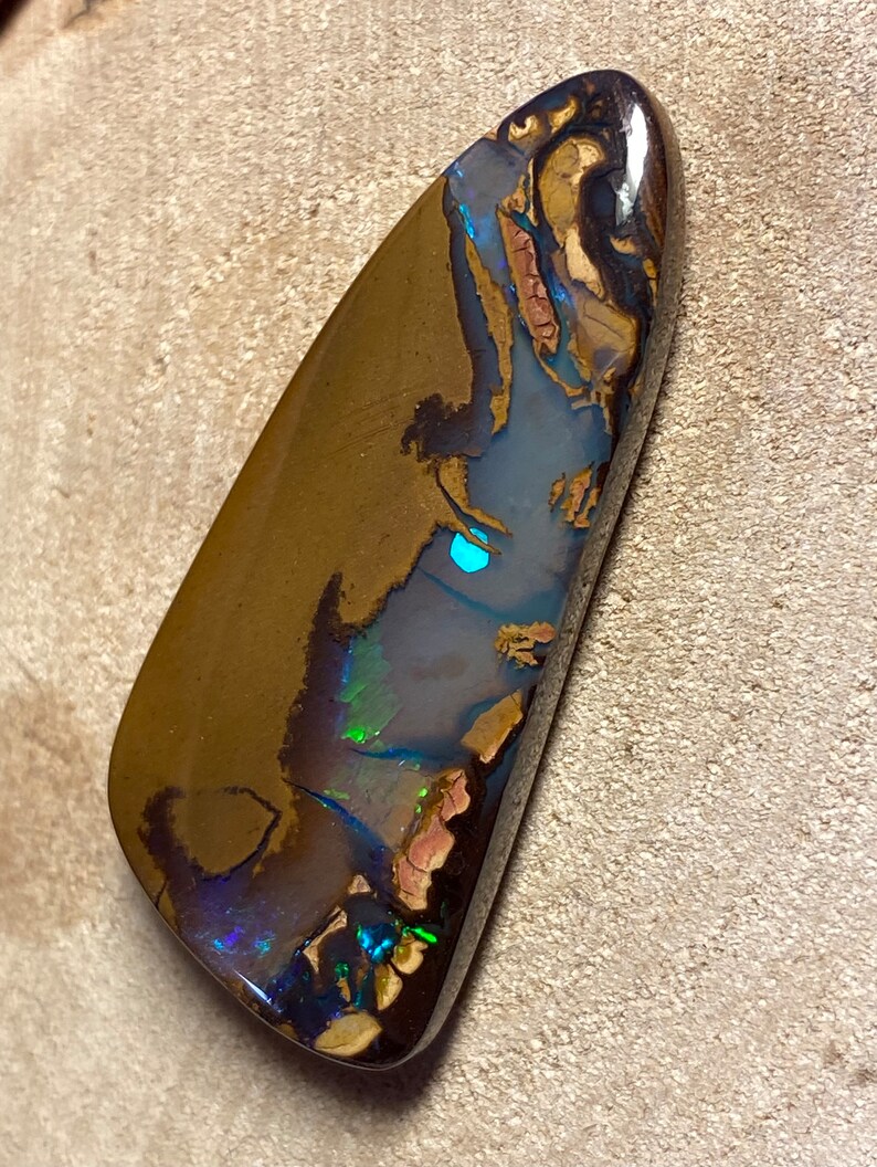 May include: A dark brown, green, and blue opal cabochon with a teardrop shape. The opal has a high polish and shows flashes of color.
