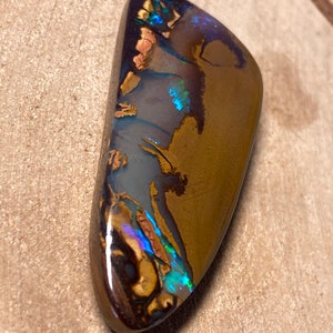 May include: A brown, iridescent opal cabochon with blue and green flashes of color. The opal is cut in a teardrop shape and has a smooth, polished surface.