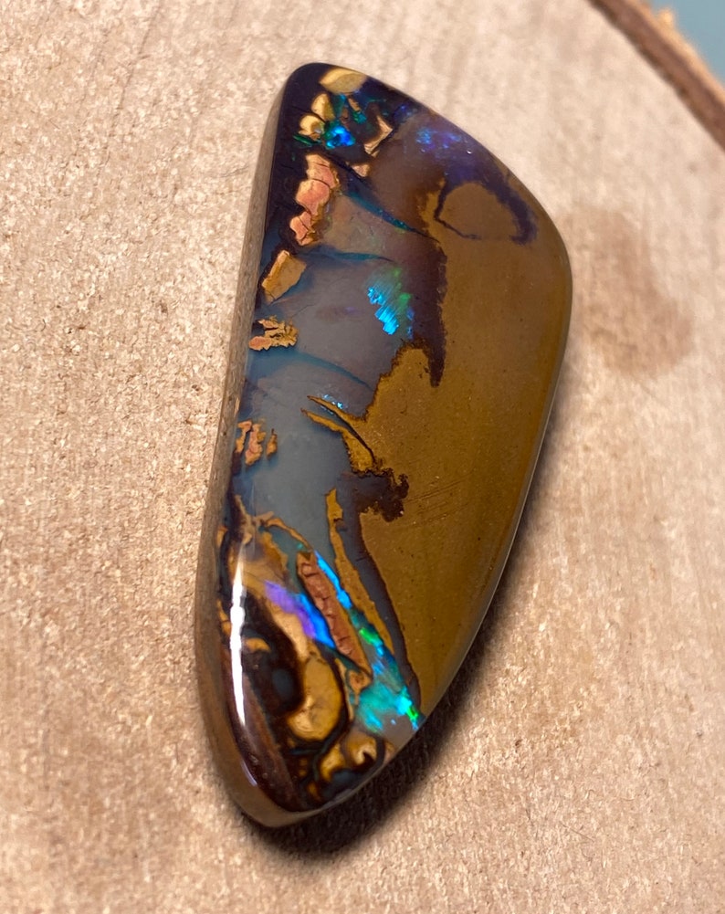 May include: A brown, iridescent opal cabochon with blue and green flashes of color. The opal is cut in a teardrop shape and has a smooth, polished surface.