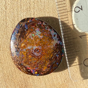 Może przedstawiać: Brązowy, opalizujący kaboszon z opalizującym wzorem w kolorze niebieskim, zielonym i czerwonym. Opal ma średnicę około 2,5 cm.