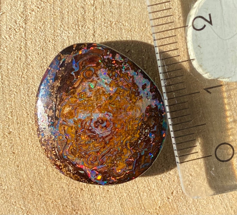 Może przedstawiać: Brązowy, opalizujący kaboszon z opalizującym wzorem w kolorze niebieskim, zielonym i czerwonym. Opal ma średnicę około 2,5 cm.