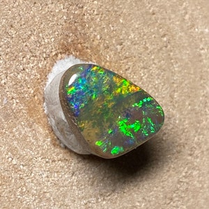 Peut inclure: Une pierre d'opale polie, de forme libre, avec un affichage de couleurs vives. L'opale présente des éclats de teintes vertes, bleues et jaunes. La pierre précieuse est posée sur une base blanche, mettant en valeur sa beauté naturelle. L'opale est une pièce unique.