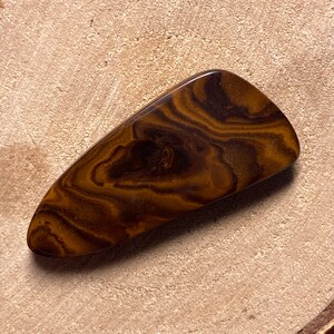 May include: A brown and gold banded agate cabochon with a smooth, polished surface. The stone has a unique, swirling pattern.