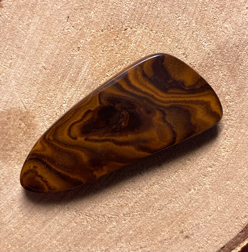 May include: A brown and gold banded agate cabochon with a smooth, polished surface. The stone has a unique, swirling pattern.