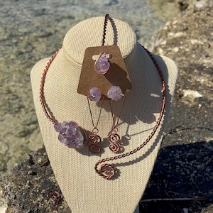 May include: A copper-toned jewelry set featuring a necklace, earrings, and a pendant. The necklace and earrings incorporate raw amethyst stones. The earrings have a spiral design. The pendant is teardrop-shaped. The jewelry is displayed on a mannequin head.