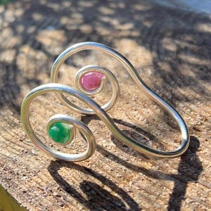 May include: A silver-toned, abstract ring with a swirling design. The ring features a pink and a green gemstone set within the swirls. The ring is displayed on a wooden surface.