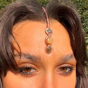 May include: A copper-colored forehead jewelry piece with three stones: a blue-green, a yellow, and a brown stone. The stones are set in a vertical line, connected by a twisted copper wire. The jewelry is worn on the forehead.