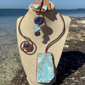 May include: A handcrafted jewelry set featuring a necklace, earrings, and a ring. The necklace has a large, rectangular, light blue stone pendant. The earrings and ring have smaller blue stones. All stones are set in copper wire.