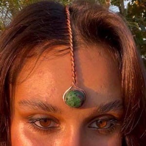 Peut inclure: Un bijou de tête cuivré avec un design torsadé, orné d'un pendentif en pierre précieuse verte et rouge. Le bijou est porté sur le front, la pierre précieuse reposant entre les sourcils. L'image est un gros plan du front et des yeux.