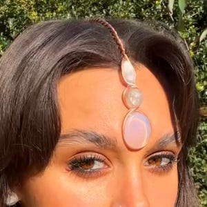 May include: A copper-toned headpiece with three oval and round gemstones. The gemstones are white, clear, and pink. The headpiece is worn on the forehead and is attached to a braided copper wire.