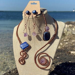 May include: A handcrafted jewelry set featuring a necklace and earrings. The necklace and earrings are made with copper wire and feature blue lapis lazuli and purple amethyst gemstones. The necklace has a spiral design and the earrings have a dangling design.