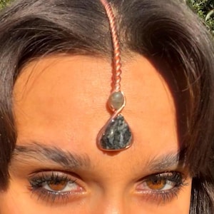 Pode incluir: Um adorno frontal cor de cobre com um pingente de pedra cinza escuro em forma de gota. O pingente é suspenso por uma corrente de cobre torcida, com uma pedra menor verde claro acima. O adorno é usado na testa.