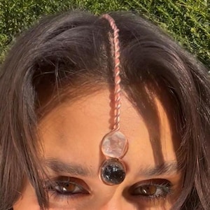 Pode incluir: Uma peça de cabeça cor de cobre com um cordão rosa torcido e duas pedras de cristal. A pedra superior é transparente e a inferior é preta. A peça de cabeça é usada na testa.