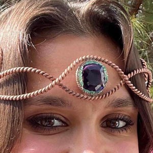 Pode incluir: Uma tiara de metal cor de cobre com uma grande pedra preciosa quadrada, roxa escura, no centro. A pedra é rodeada por uma borda verde clara. A tiara tem um design de corda torcida.