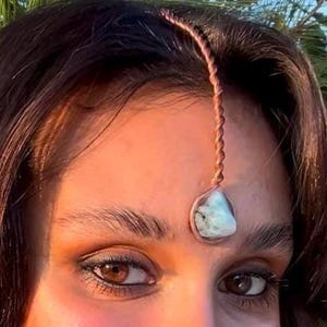 Pode incluir: Um adorno frontal cor de cobre com uma corrente torcida e uma pedra preciosa azul clara. A joia é usada na testa, com a pedra preciosa repousando entre as sobrancelhas. A pessoa tem olhos castanhos e cabelo escuro.