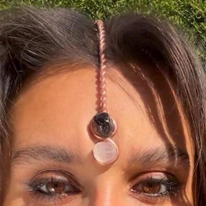 Pode incluir: Um acessório de cabelo cor de cobre com um design trançado, apresentando uma pedra preta e uma pedra branca. O acessório é usado na testa, com as pedras repousando entre as sobrancelhas.