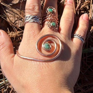 Peut inclure: Une main ornée de bijoux. On distingue une bague argentée tressée, deux bagues en fil de cuivre avec des pierres vertes et une autre bague argentée tressée. Un bracelet spirale en cuivre avec une pierre verte est également visible.