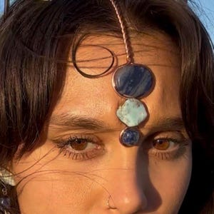 Pode incluir: Uma joia de testa em tom de cobre com três pedras preciosas: uma pedra grande, oval, azul escuro, uma pedra azul clara de formato irregular e uma pedra pequena, azul escuro. As pedras são conectadas por uma fina corrente de cobre.