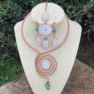 May include: A handcrafted copper wire jewelry set featuring a necklace and earrings. The necklace has a spiral pendant and a choker design. The earrings and necklace feature rose quartz, green stones, and white stones. The jewelry is displayed on a beige mannequin head.