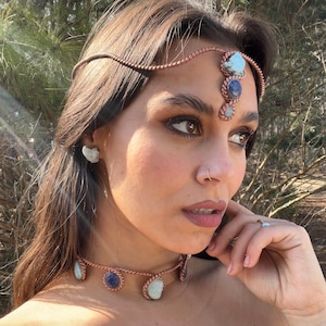 May include: A copper-toned headpiece, choker, and earrings set with blue and white gemstones. The headpiece features a braided design with stones at the forehead. The choker and earrings have similar stone accents. The jewelry is worn by a person.