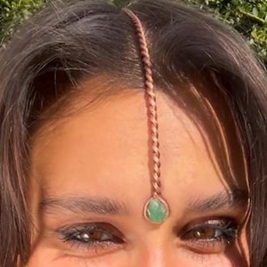 May include: A copper-colored forehead jewelry piece with a braided chain design. The chain descends from the hairline to a small, oval, green gemstone centered between the eyebrows. The jewelry is worn on the forehead.