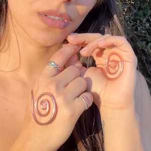Puede incluir: Dos anillos en espiral de cobre, cada uno con un pequeño centro de piedra rosa, se muestran en las manos de una persona. Los anillos son un diseño único y artesanal. La persona también lleva dos anillos de plata en los dedos.