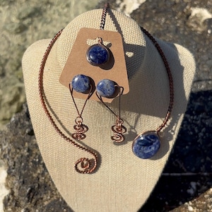 May include: A jewelry set featuring a necklace, earrings, and a pendant. The set includes a copper-colored wire necklace with a blue stone pendant, matching earrings, and a pendant. The earrings have a unique wire design. The jewelry is displayed on a beige mannequin head.