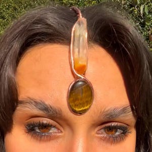 Pode incluir: Uma peça de cabeça com fio de cobre com um cristal de quartzo transparente e uma pedra olho de tigre. A peça de cabeça é projetada para ser usada na testa. A pedra olho de tigre oval tem uma cor marrom dourado.
