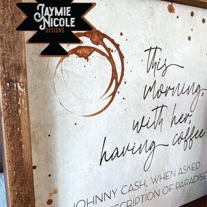 May include: A white wooden sign with brown wood framing. The sign has a coffee stain design and the text "This morning, with her having coffee - Johnny Cash, when asked for his description of paradise."