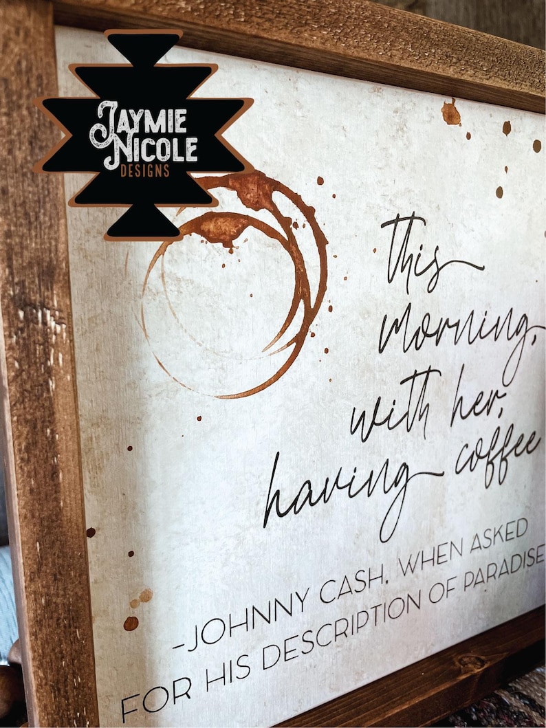 May include: A white wooden sign with brown wood framing. The sign has a coffee stain design and the text "This morning, with her having coffee - Johnny Cash, when asked for his description of paradise."