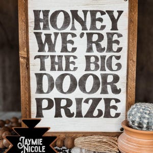 Puede incluir: Cartel enmarcado en madera con el texto "HONEY WE'RE THE BIG DOOR PRIZE" en negro sobre fondo blanco. El cartel tiene un marco de madera marrón. También se ve un pequeño cactus en una maceta de terracota.