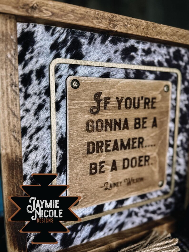 May include: Wooden framed sign with a cowhide pattern background. The sign features a wooden plaque with the quote "If you're gonna be a dreamer... be a doer." The sign also includes the text "Jaymie Nicole Designs."