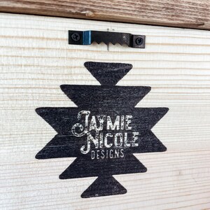 May include: A wooden panel featuring a black geometric design and the text "Jaymie Nicole Designs" in white script. A metal sawtooth hanger is attached to the top. The wood has a natural grain and a light brown border.