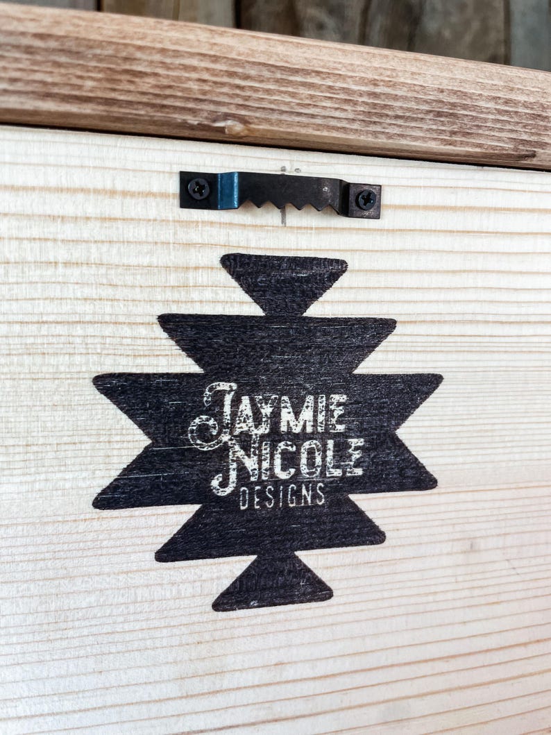 May include: A wooden panel featuring a black geometric design and the text "Jaymie Nicole Designs" in white script. A metal sawtooth hanger is attached to the top. The wood has a natural grain and a light brown border.