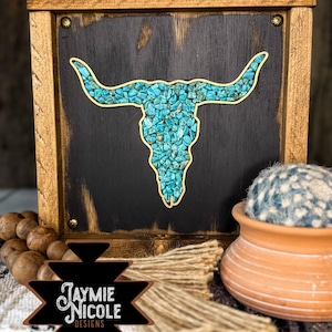 May include: A wooden framed wall art piece featuring a bull skull design. The skull is filled with turquoise-coloured stones and outlined in gold. The background is black, and the frame is a light brown colour. The artwork is branded with the text "Jaymie Nicole Designs."