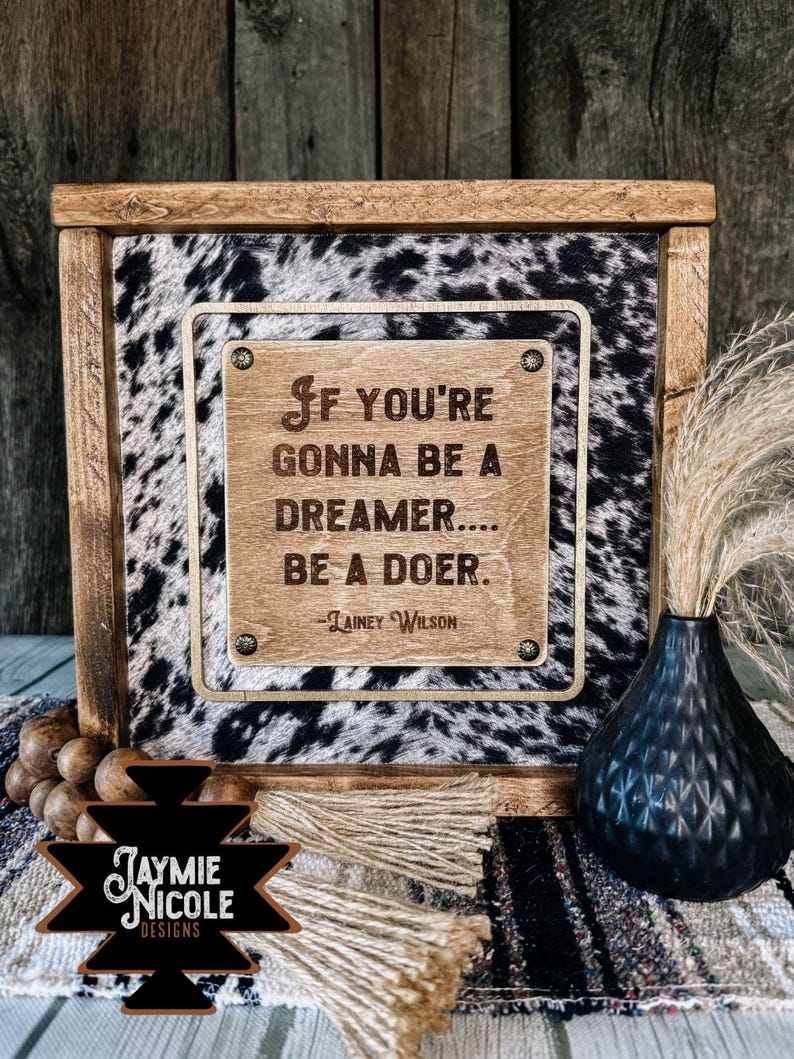 May include: A framed sign with a cowhide pattern and a wooden plaque bearing the quote "If you're gonna be a dreamer... be a doer." The sign also features the text "Jaymie Nicole Designs."