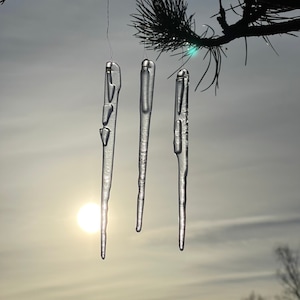 以下が含まれることがあります： 曇り空に、松の枝からぶら下がっている透明なガラスのつらら3つ。背景には太陽が輝いています。
