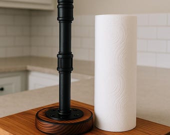 Puede incluir: Un portarrollos de papel de cocina de metal negro y madera con un rollo de papel blanco. El soporte tiene una base redonda de madera teñida y una estructura negra similar a una tubería. Las toallas de papel son blancas con un patrón sutil.
