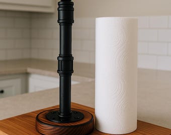 Puede incluir: Un portarrollos de papel de cocina de metal negro y madera con un rollo de papel blanco. El soporte tiene una base redonda de madera teñida y una estructura negra similar a una tubería. El rollo de papel de cocina es blanco y cilíndrico, sobre una tabla de cortar de madera.