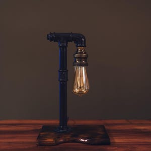 Könnte beinhalten: Eine schwarze Schreibtischlampe im Rohrstil mit einer Vintage-Edison-Glühbirne. Die Lampe ist auf einem dunklen Holzsockel montiert. Die Metallkomponenten der Lampe sind schwarz, und die Glühbirne strahlt ein warmes, goldenes Licht aus. Das Design ist industriell und rustikal.