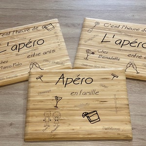 May include: Three wooden chopping boards with laser-engraved designs. The boards feature the text "C'est l'heure de L'apéro" and various drinks and locations. The middle board says "Apéro en famille" with a couple and a cocktail.