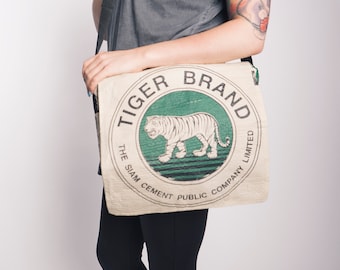 May include: A beige canvas messenger bag with a black strap. The bag features a circular logo with a green background and a white tiger. The logo reads "Tiger Brand The Siam Cement Public Company Limited".