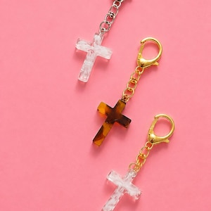 May include: Three cross charms with metal lobster claw clasps and short chains. Two charms feature a white marbled pattern with silver and gold hardware, while the center charm has a brown tortoiseshell pattern with gold-toned hardware.